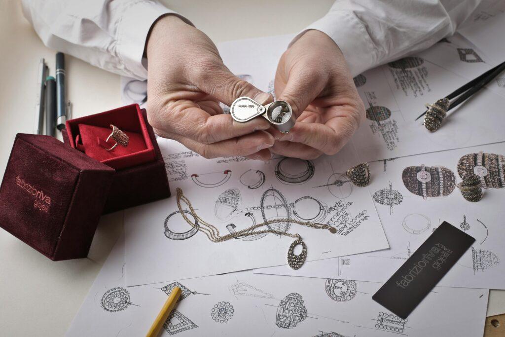 pexels-photo-1050321-1050321 Close-up of hands inspecting a diamond ring with a loupe on a workbench filled with jewelry sketches.